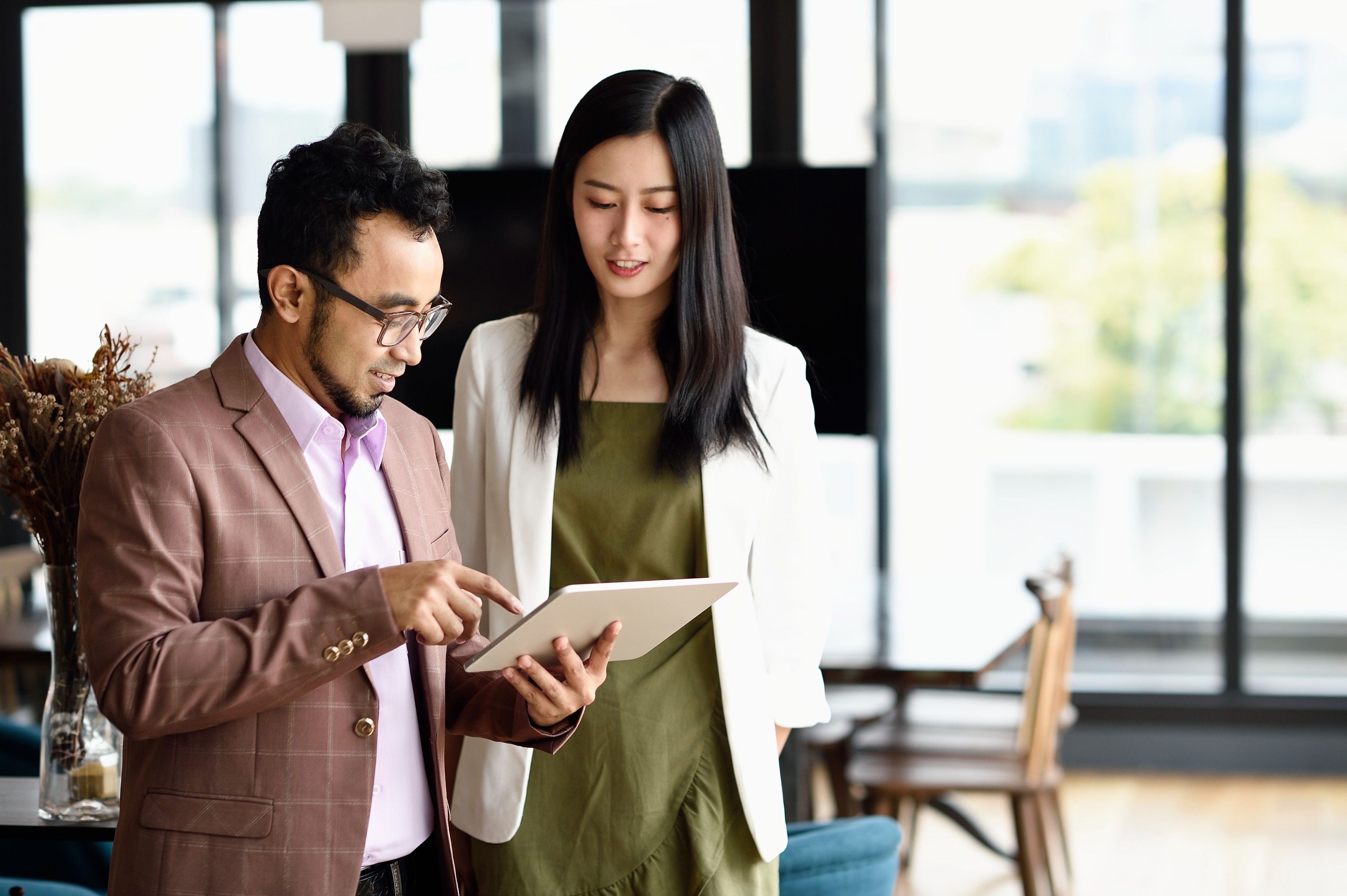 Two people discuss collaboration in an open plan office space while looking at a tablet device.