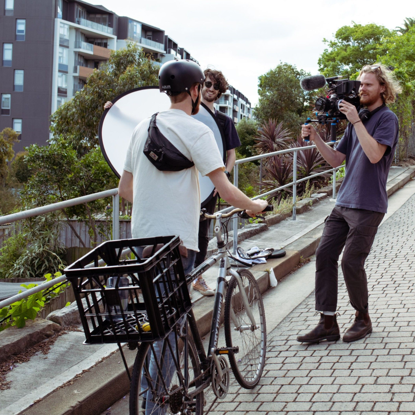 A person on a bicycle interacts with a solopreneur filmmaker holding a camera, while another person holds a reflector.
