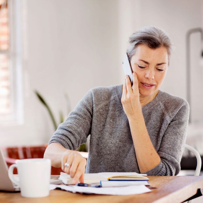 A busy person talks on the phone while sitting in front of a messy desk in their kitchen.