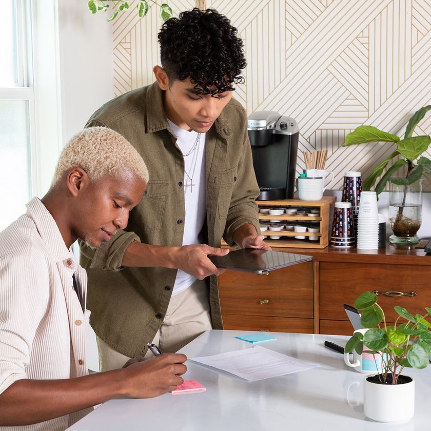 A man sat at a table writing on a post-it note whilst being instructed by a man reading off of a tablet.