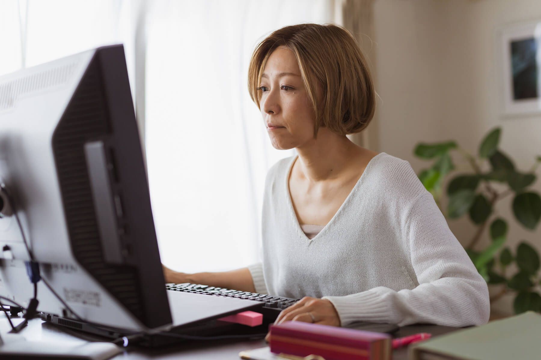 A woman sits at a desk at home, looking intently at the computer screen