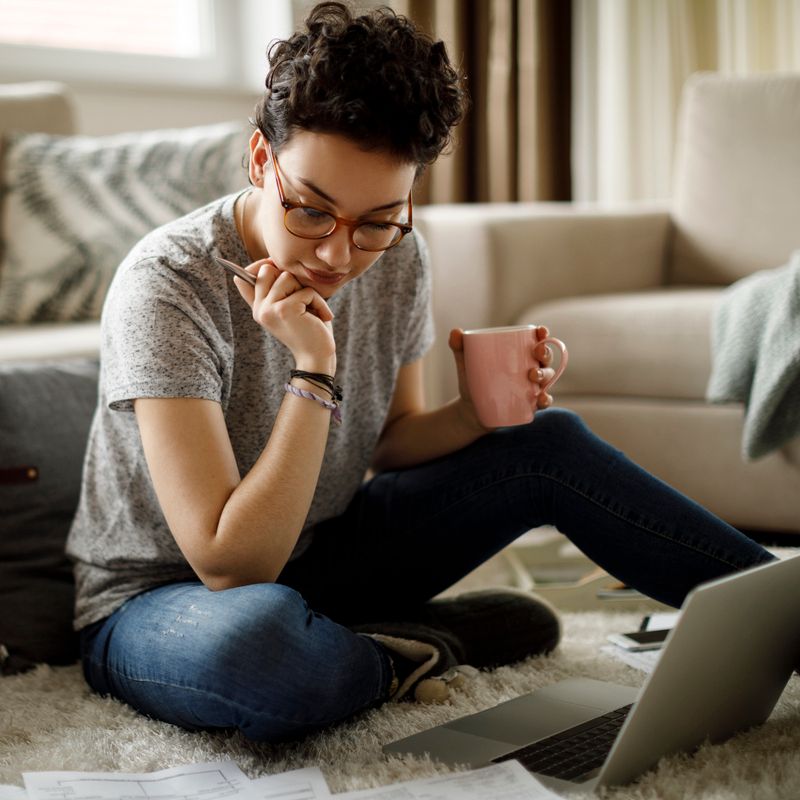 A woman sits on the floor surrounded by papers and looks at her laptop with a mug in hand.