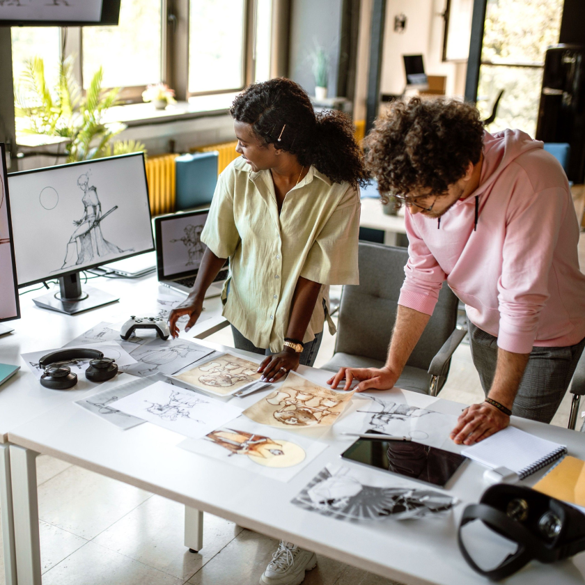 Two creative professionals lean forward on a desk, looking at drawings on a table and a concept on a computer monitor.