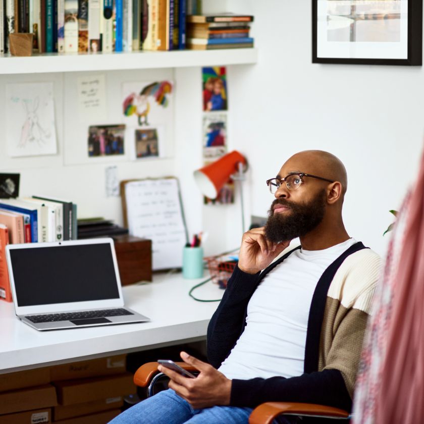 Man sits thinking at desk in home office surrounded by books and laptop.
