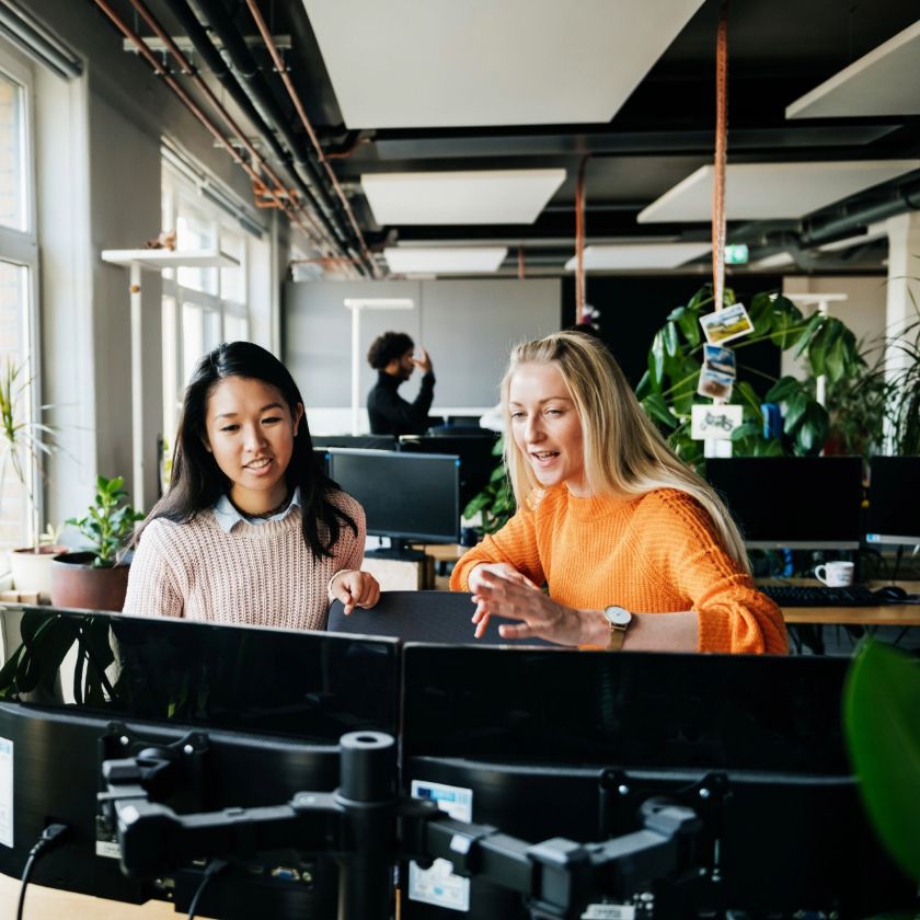 Two people sit at a desk in an open plan workspace while discussing file sharing apps.