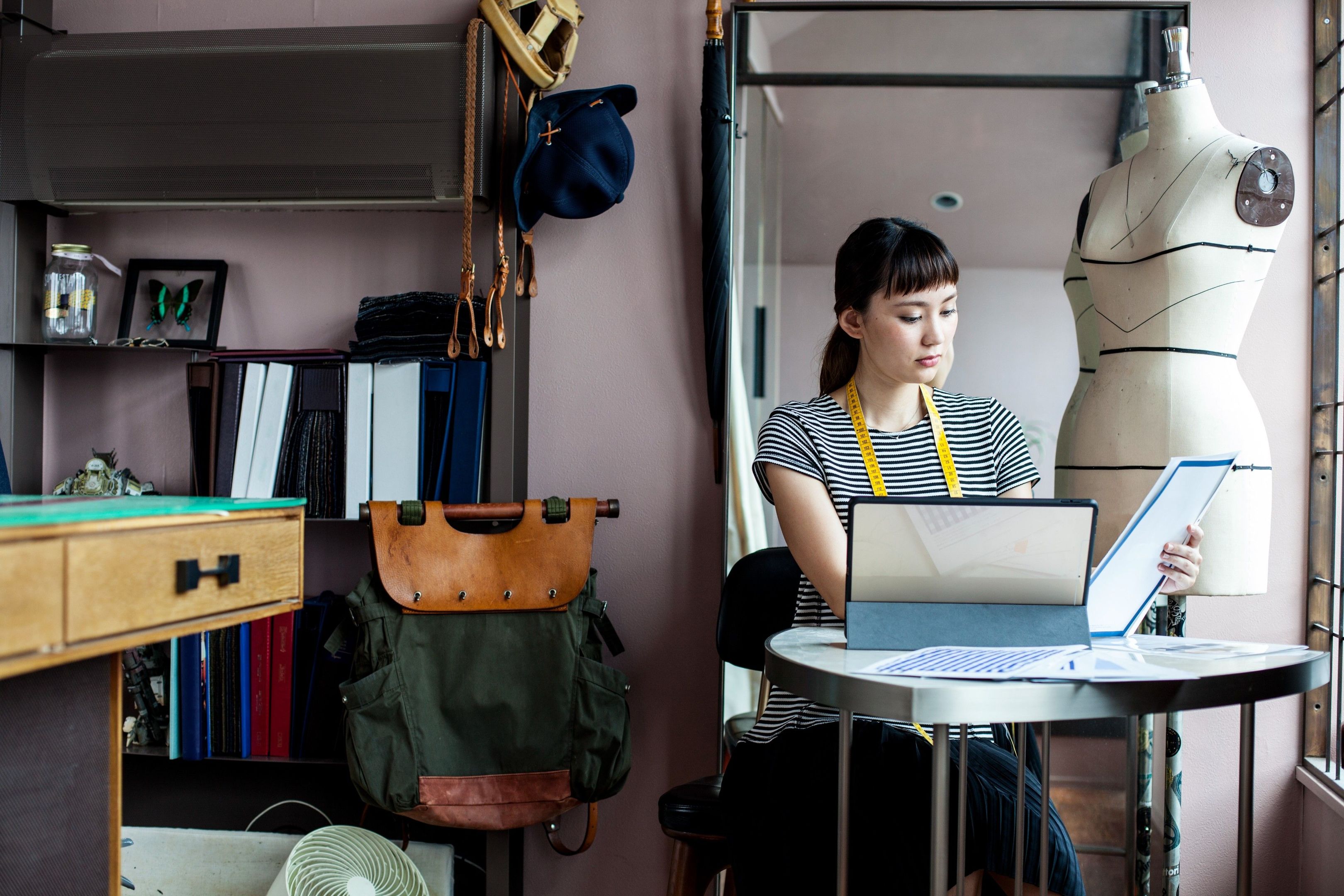 A woman sits at a small table with a tablet whilst looking at notes/documents/papers, with a tape measure draped around her neck. Behind her is a clothes mannequin suggesting a creative profession.