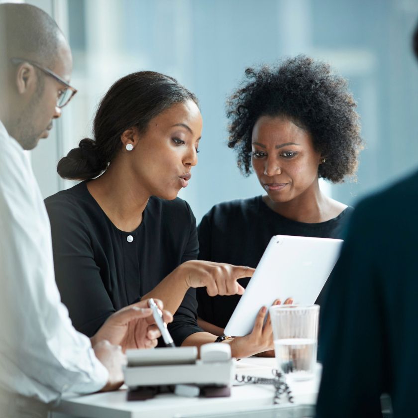 A group of people having a meeting in a modern office, one person points at a tablet device while others sit around a table.