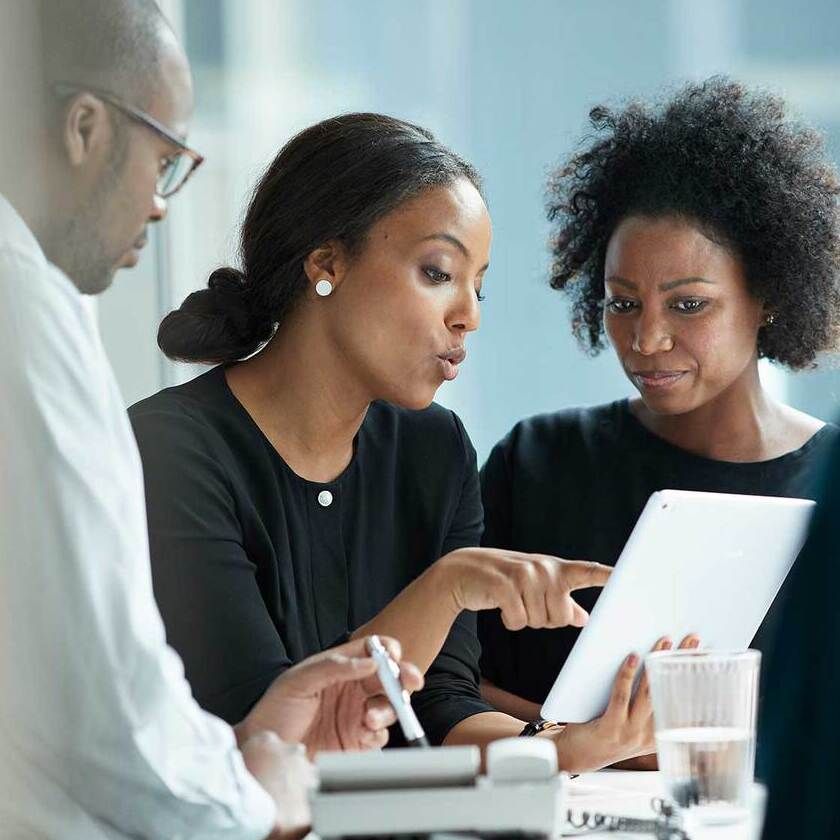 A group of people in an office meeting, with two women discussing their work on an ipad.