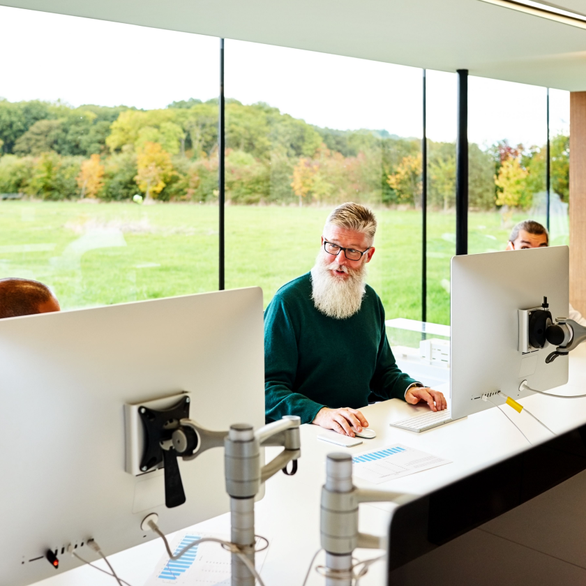 Older man with a white beard working at a desk with large computer monitors in a bright office with floor-to-ceiling windows overlooking a green landscape. He is engaged in conversation with colleagues seated nearby.