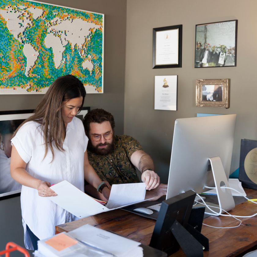 Two people at a desk, one sitting, one standing, review a script and a video to provide feedback.