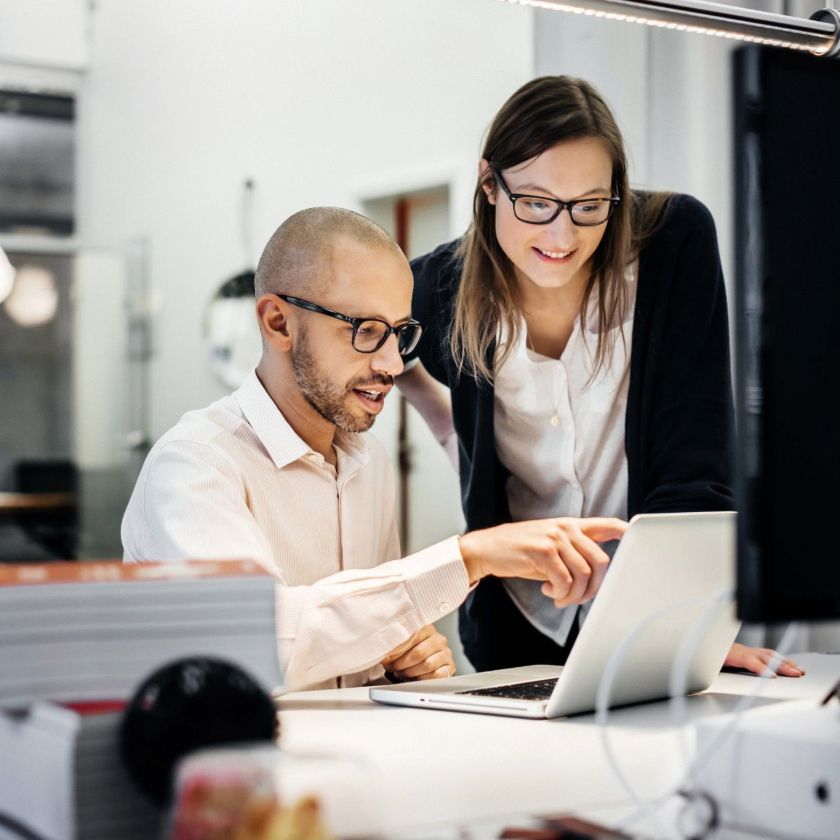 Two people collaborate at a desk, reviewing content on a laptop in a modern office setting.