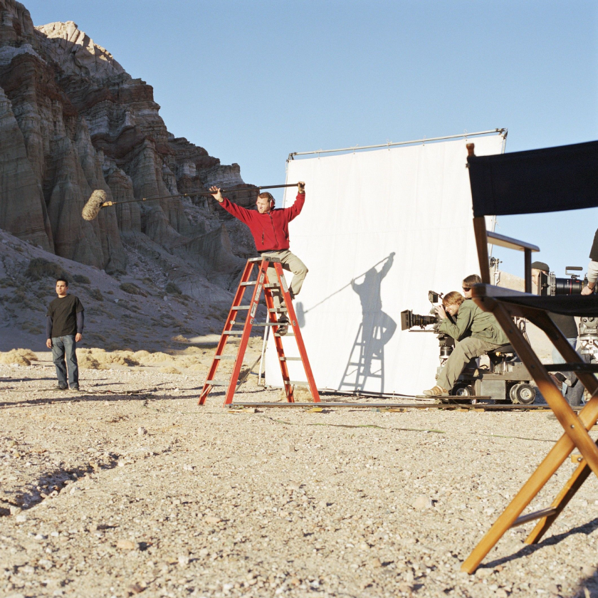 Film crew on a desert set with a white screen backdrop. A person on a ladder holds a boom mic while a camera operator films.