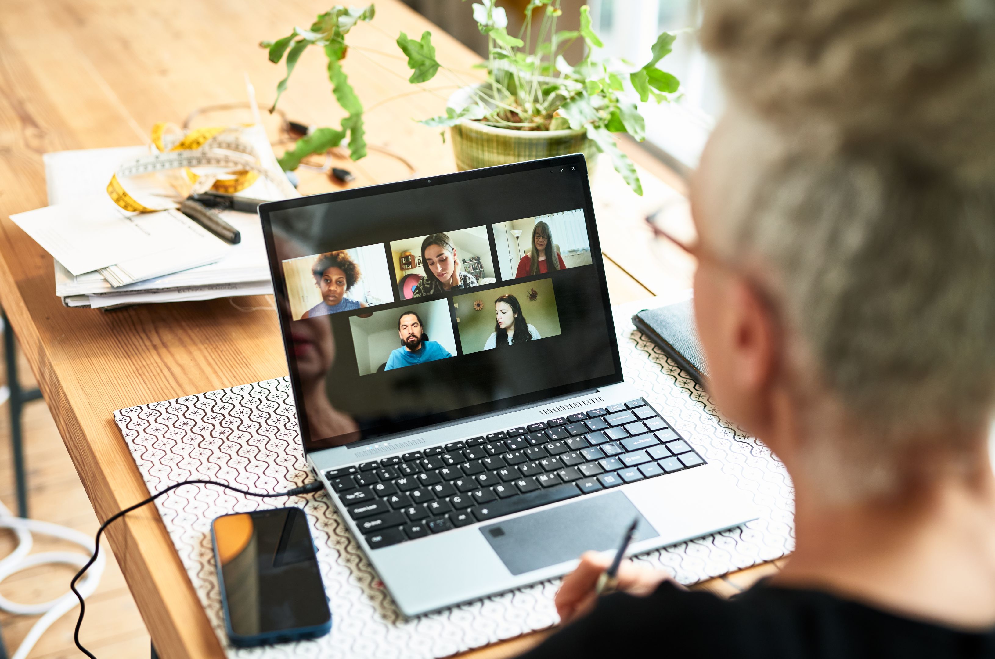 Looking over a remote worker's shoulder, a laptop sits open with five team members on a video call discussing a project.