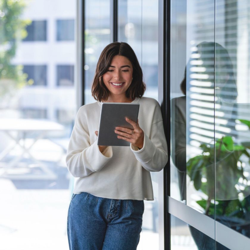 A person stands outside a building, leaning against a glass wall while looking at a contract on their tablet.