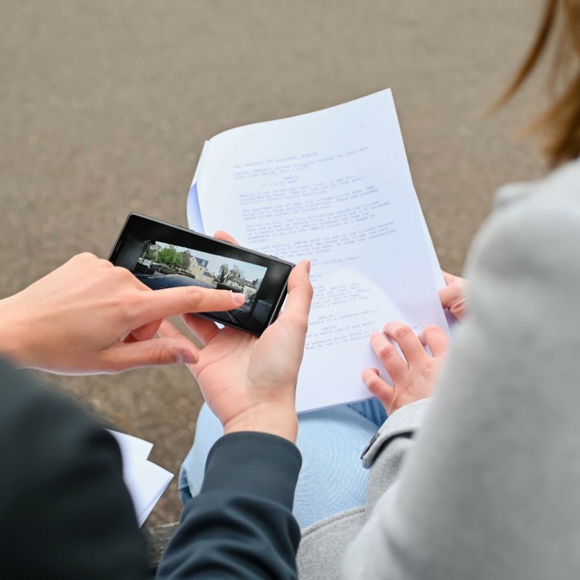 An over-the-shoulder view of a person holding a phone to review footage, while another consults a script.