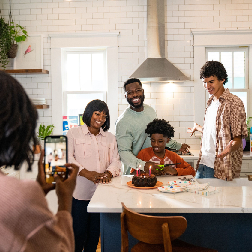 Using a mobile device, someone takes a photo of a family standing around a child and their birthday cake.