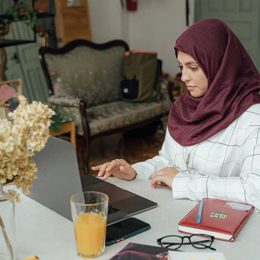 Person working from home at a tidy table setup with a laptop, notebook, glasses, and a glass of juice in a cozy living room.