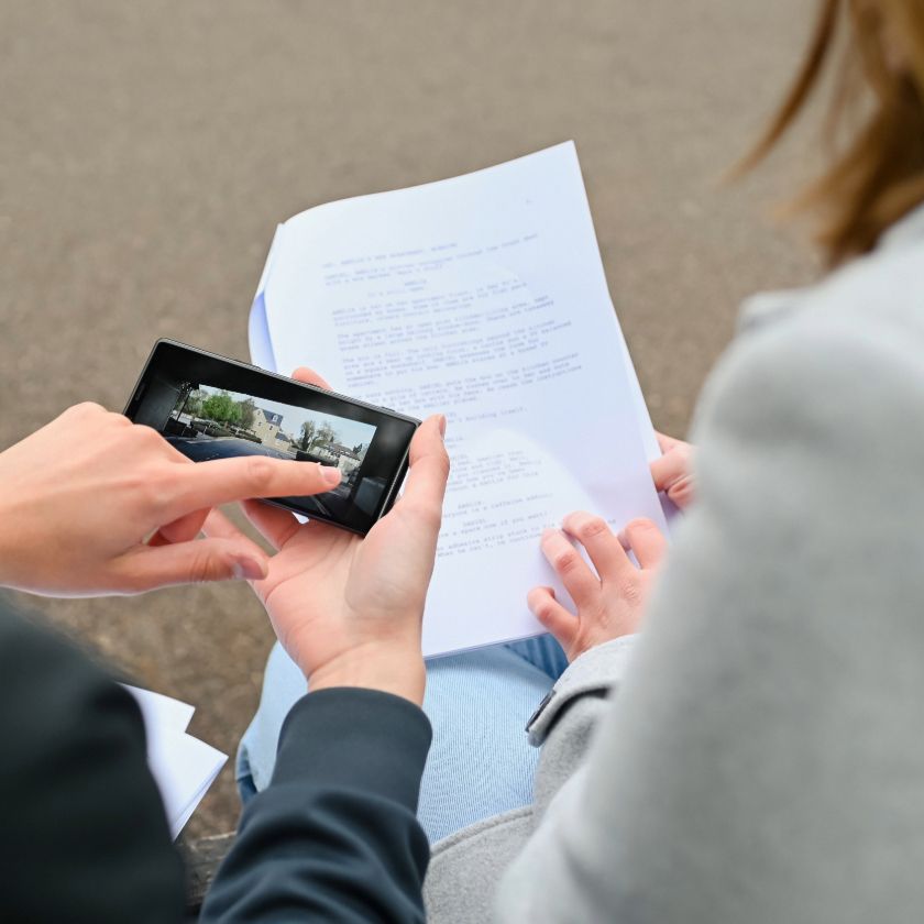 An over-the-shoulder view of a person holding a phone to review footage, while another consults a script.