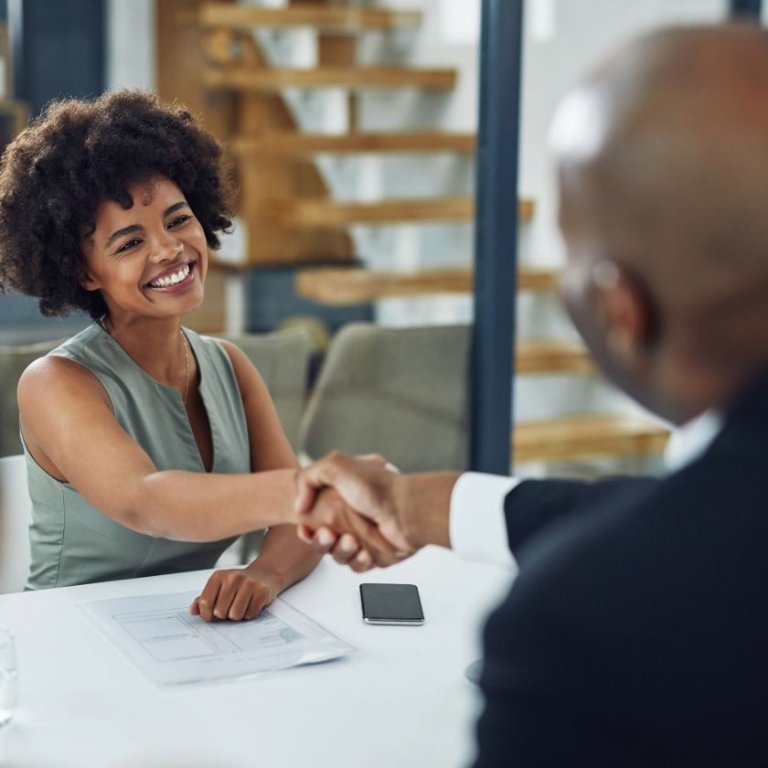 A person smiles and shakes hands with another person across a desk in an office meeting room.