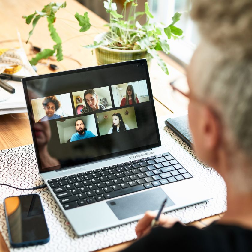 Looking over a remote worker's shoulder, a laptop sits open with five team members on a video call discussing a project.