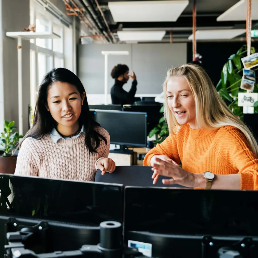 Two colleagues discussing work at dual monitors in a modern office, symbolizing collaboration and teamwork.