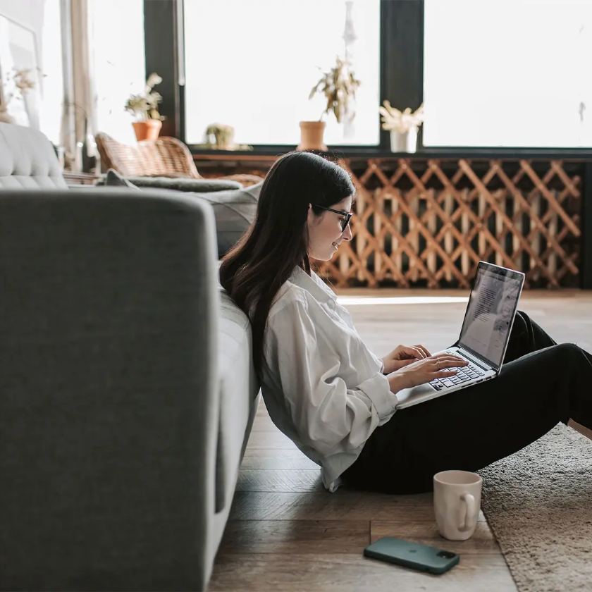 A person working remotely sits on the floor at home, leaning back against a couch as they type on their laptop.