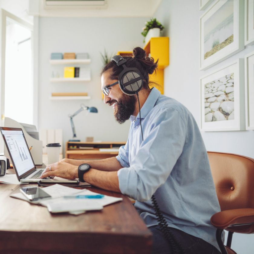 A person works from home at a desk, smiling at the laptop in front of them as they type and share files with their team.