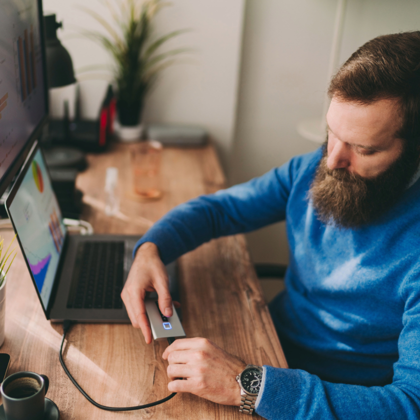 A person sat at a desk connects their external hard drive to their laptop using a USB cable to back up their files offline.