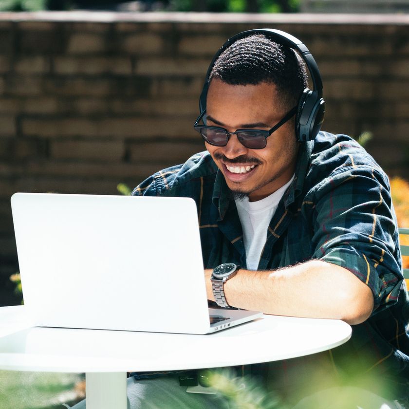 A man sits outside in the sun on his laptop, he is wearing headphone, sunglasses and is smiling widely at the screen