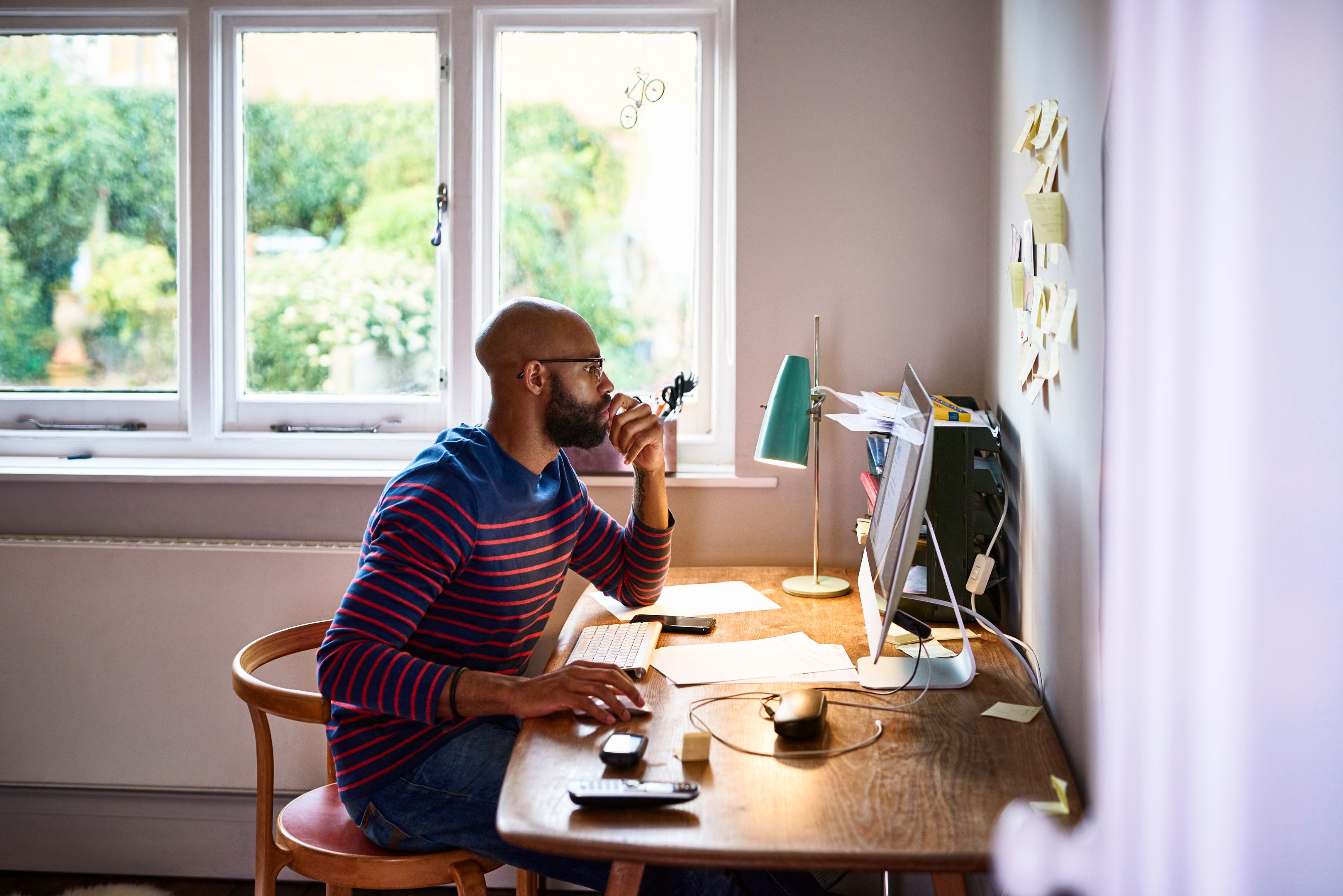A person working at home office computer, reading PDF content onscreen with documents and stationery spread across desk.