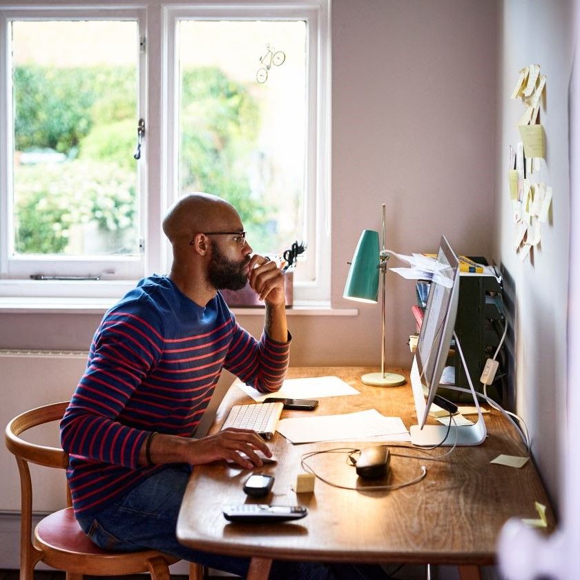 A person working at home office computer, reading PDF content onscreen with documents and stationery spread across desk.