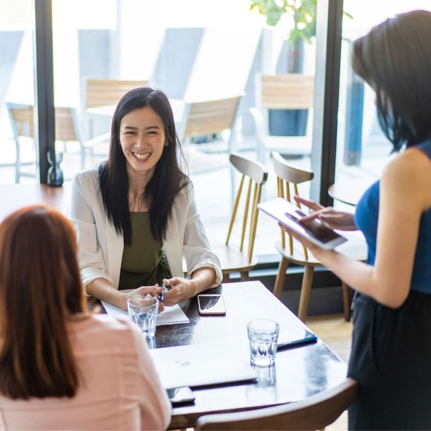 A group of workers smile as they sit around a table working together, led by their team leader using a tablet