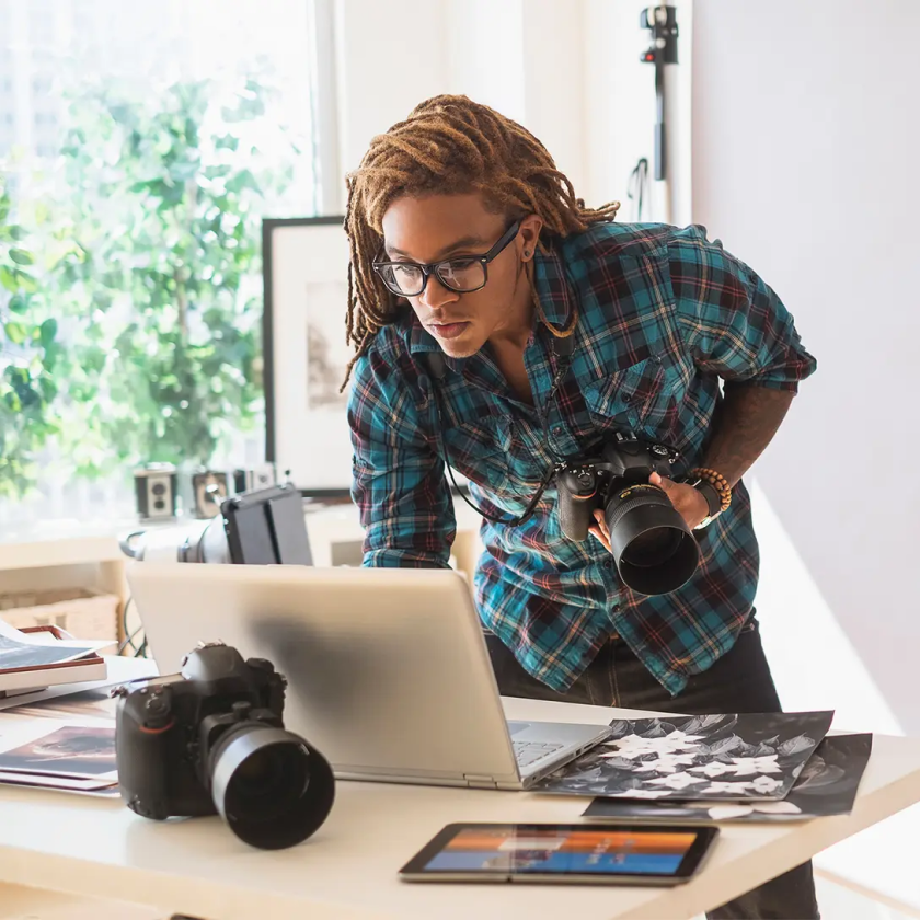 A person in a photography studio leans over their laptop while holding a professional camera.