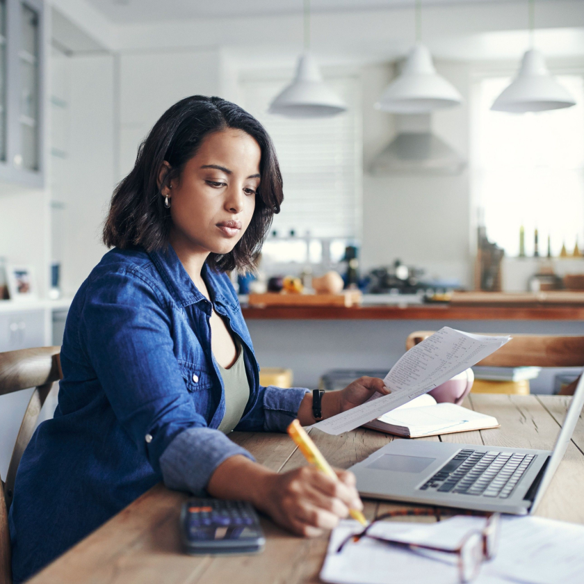 A solopreneur writes down notes on a notepad next to their laptop while holding a document in the other hand.