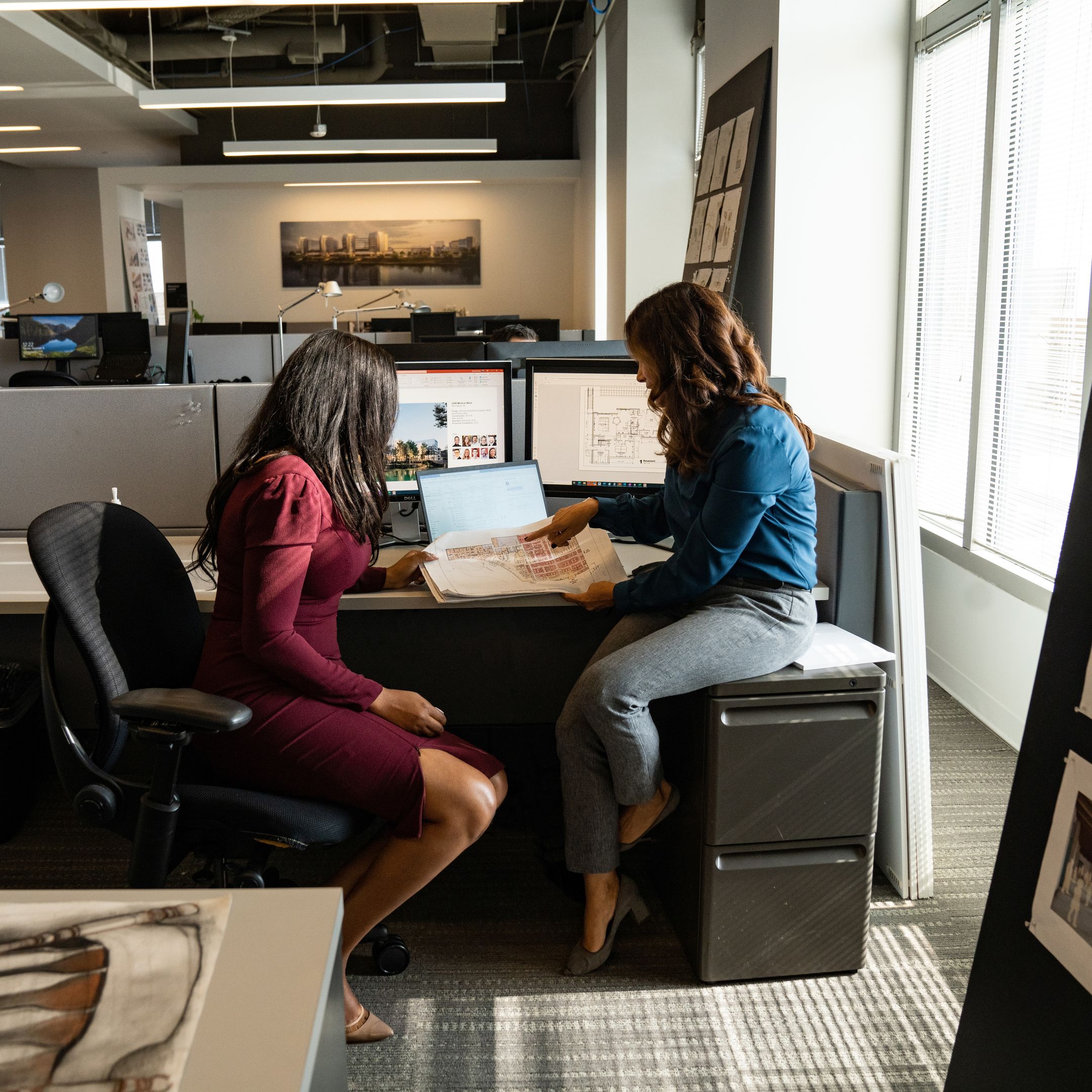 Two people sit at a desk in an open plan office space having a discussion.