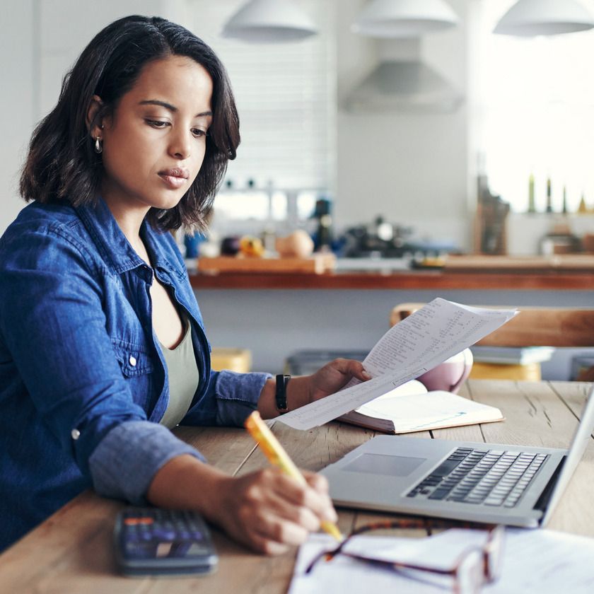 A woman sits at a table taking notes whilst browsing on her laptop, she is surrounded by documents, a planner and calculator
