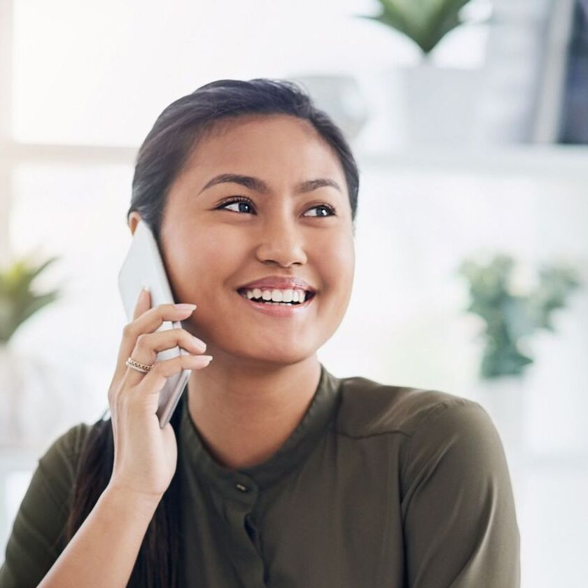 A person smiling as they sit at their laptop and enjoy a phone conversation.