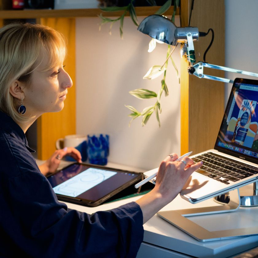 Person sitting at a desk using a digital tablet and stylus while referencing artwork on a laptop screen. The workspace is well-lit with a desk lamp, and the individual appears focused on a digital illustration project.