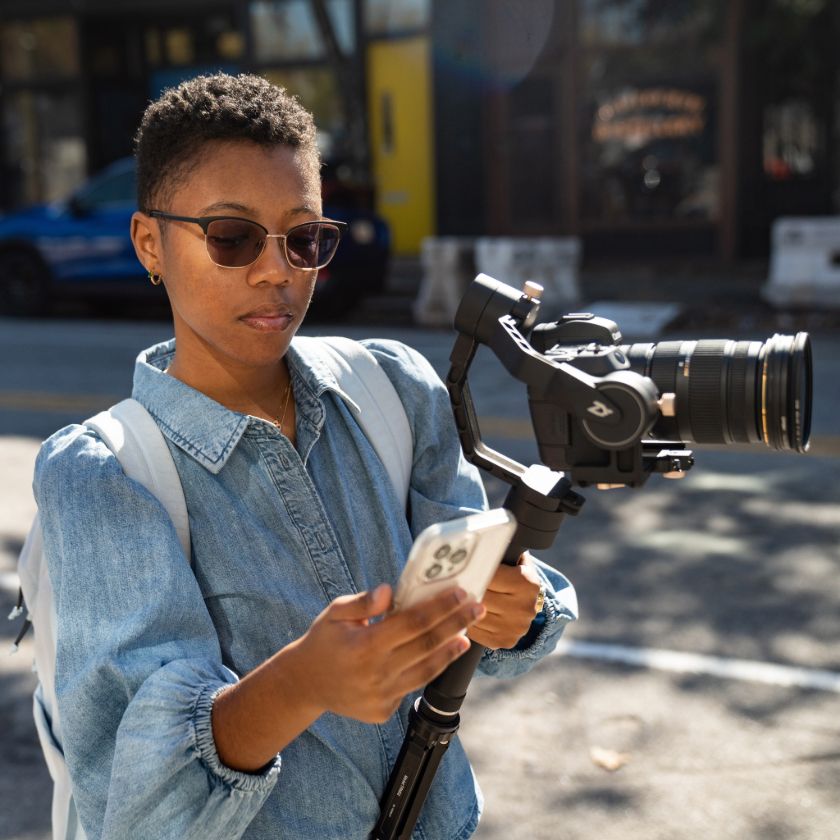 A videographer holds a camera in one hand and their phone in the other.