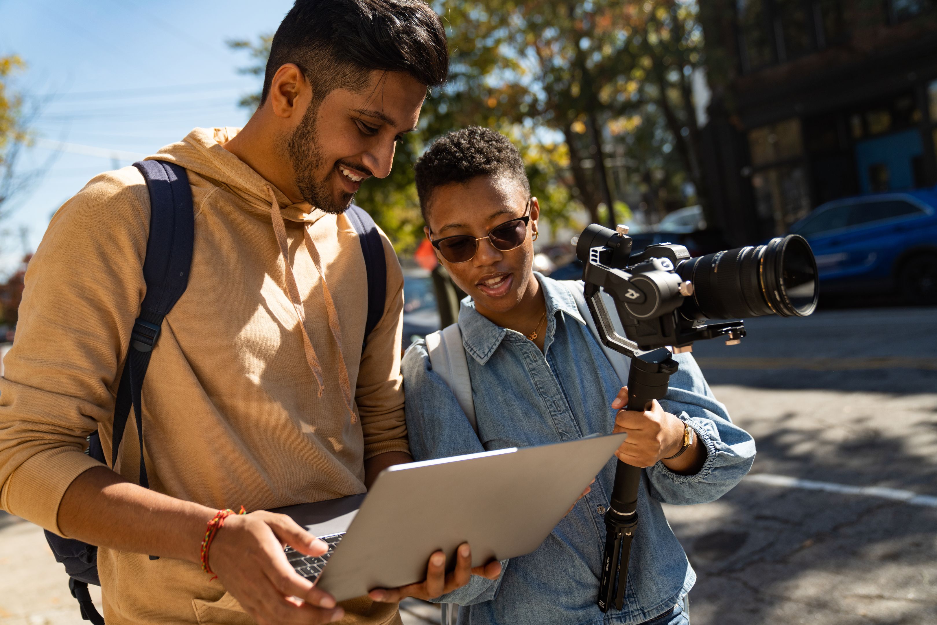 Two people stand outdoors, one holding a camera stabilizer and the other holding a laptop, both looking at the laptop screen.