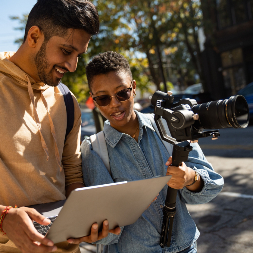 Two creatives working on a video shoot—one carrying a camera on a gimbal—check the storyboard for a shoot on an open laptop.