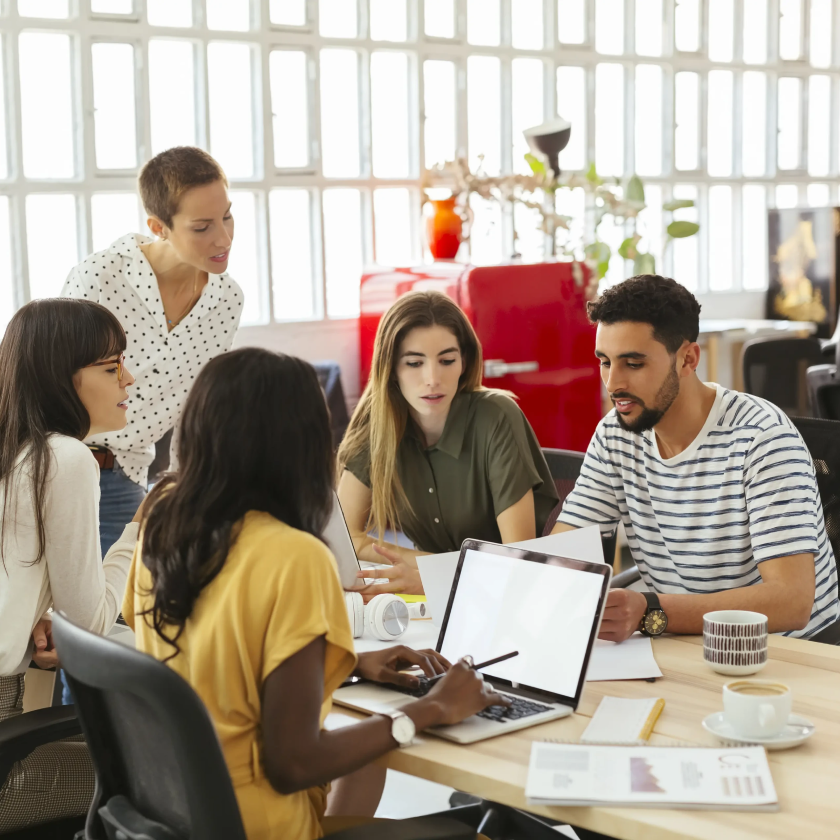 A group of people gather 'round a laptop in their physical office space, preparing for a pitch.