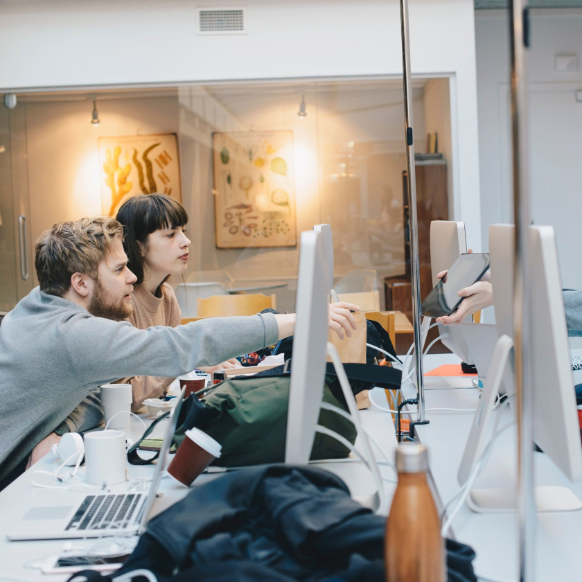 Group of people sitting at a table with computers and a tablet, engaging in a shared workspace.