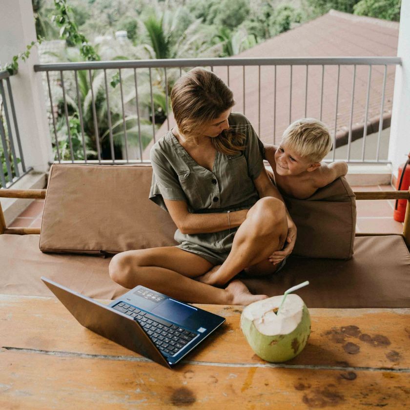A mother faces their child as they sit on a balcony using a laptop to telecommute.