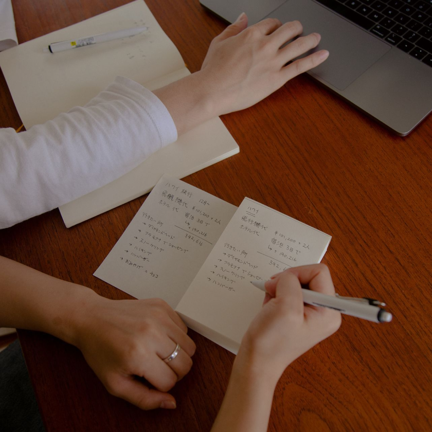 Two people sit at desk, only their hands showing as they each write in notebooks and one uses a laptop's trackpad.